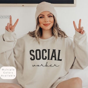 Puede incluir: Una sudadera beige con el texto "SOCIAL worker" impreso en la parte delantera. La sudadera la lleva puesta una persona con una boina beige.