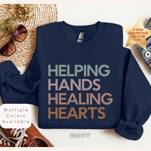 Puede incluir: Sudadera azul marino con el texto "HELPING HANDS HEALING HEARTS" en letras coloridas y superpuestas.