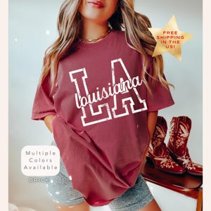May include: Brick-colored t-shirt with "LA Louisiana" in white lettering. The shirt has a crew neck and short sleeves. The image also shows a gold star with the text "FREE SHIPPING IN THE US!" and a pair of red cowboy boots.