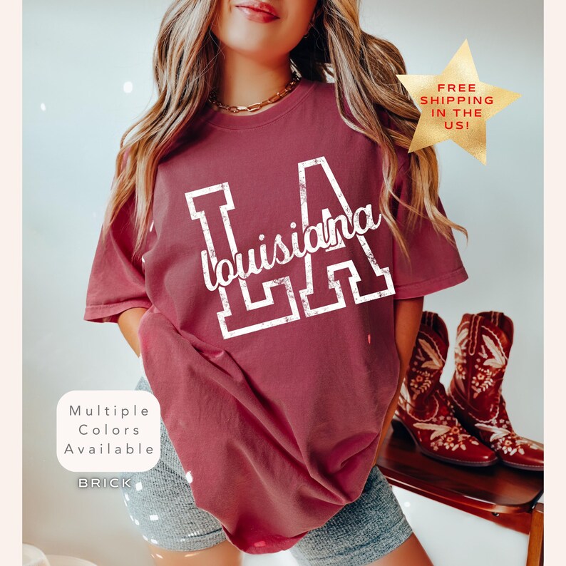 May include: Brick-colored t-shirt with "LA Louisiana" in white lettering. The shirt has a crew neck and short sleeves. The image also shows a gold star with the text "FREE SHIPPING IN THE US!" and a pair of red cowboy boots.