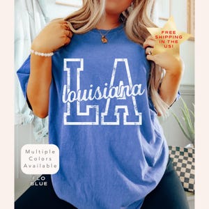 May include: A blue t-shirt with the text "LA Louisiana" in white block letters and script. The shirt has a relaxed fit and is labeled "Flo Blue". The image also includes a gold star with the text "FREE SHIPPING IN THE US!"