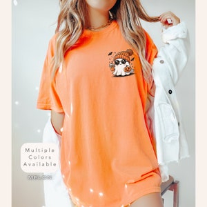 Pode incluir: Uma camiseta laranja brilhante com um fantasma de desenho animado fofo usando um gorro e segurando uma bebida em forma de ab&oacute;bora. O fantasma usa &oacute;culos de sol e um pequeno sorriso. O texto "Multiple Colors Available" e "MELON" est&atilde;o impressos abaixo da imagem.