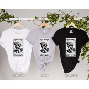 May include: Three t-shirts in white, ash grey, and black with a graphic of a skeleton wearing a beanie and eating a pickle. The graphic is surrounded by a rectangle with the text "Dead Serious About Pickles".