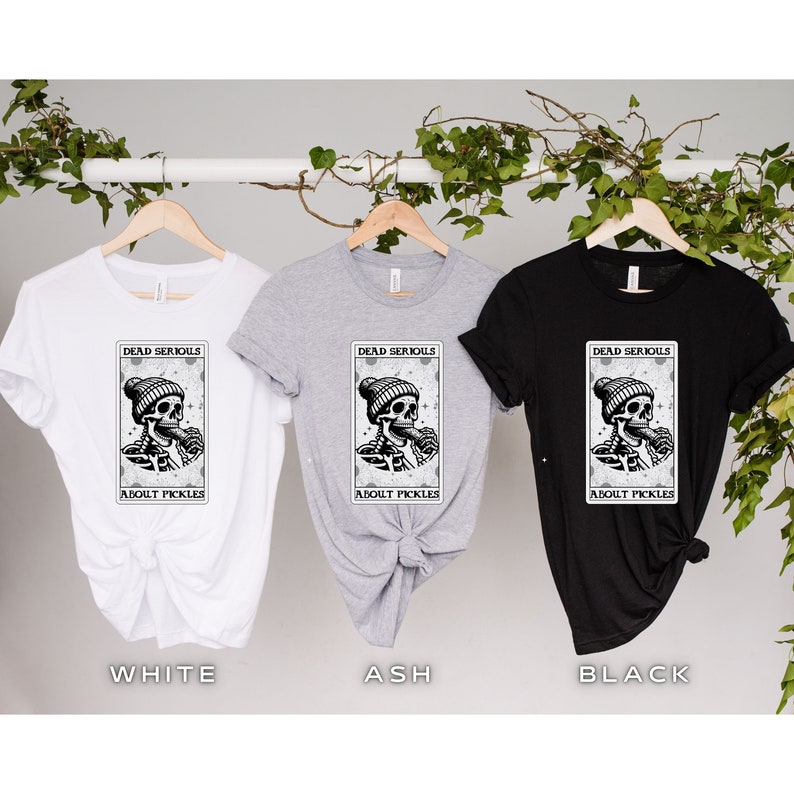 May include: Three t-shirts in white, ash grey, and black with a graphic of a skeleton wearing a beanie and eating a pickle. The graphic is surrounded by a rectangle with the text "Dead Serious About Pickles".