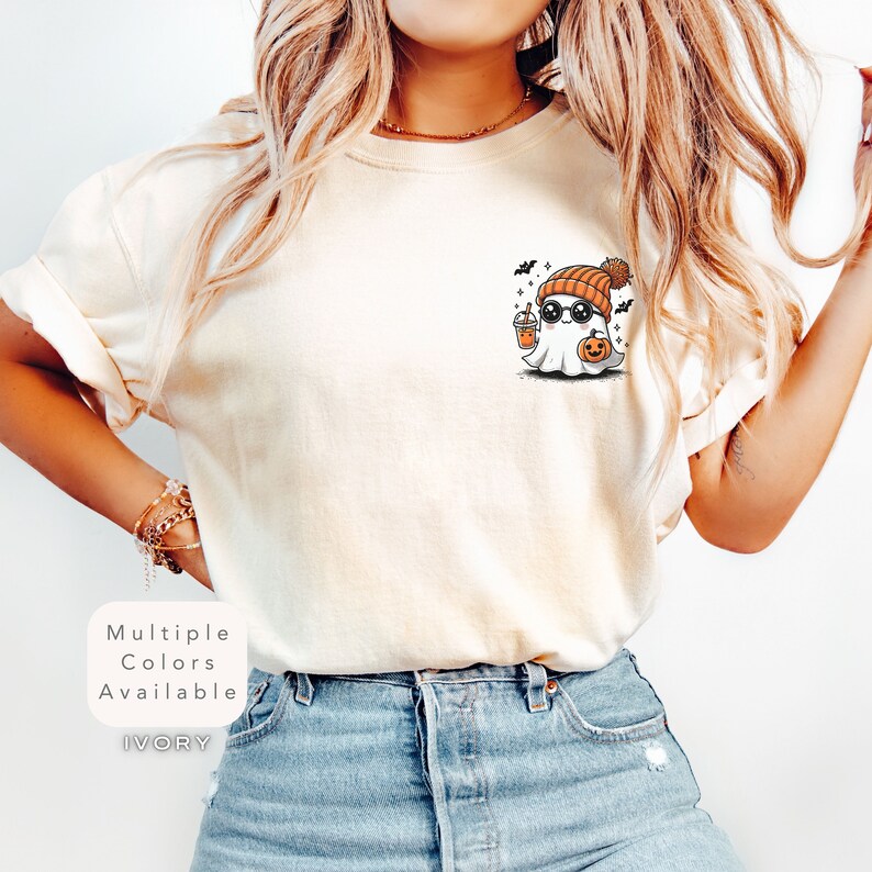Pode incluir: Uma camiseta branca com um fantasma de desenho animado fofo usando um gorro e &oacute;culos de sol, segurando uma ab&oacute;bora e uma x&iacute;cara de caf&eacute;. O texto "Multiple Colors Available" e "IVORY" est&aacute; impresso na camiseta.