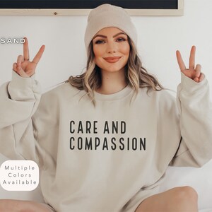 Puede incluir: Una sudadera beige con el texto "CARE AND COMPASSION" impreso en negro. La modelo lleva la sudadera y una boina beige.