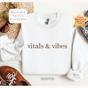 May include: White crewneck sweatshirt with the text "vitals & vibes" printed in brown on the front. The sweatshirt is laying flat on a white surface.