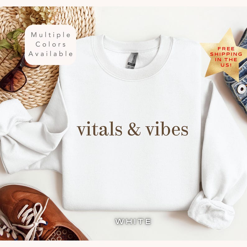 May include: White crewneck sweatshirt with the text "vitals & vibes" printed in brown on the front. The sweatshirt is laying flat on a white surface.