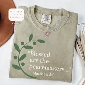 May include: A khaki green t-shirt with a white graphic print of a leafy branch and the text "Blessed are the peacemakers... - Matthew 5:9".