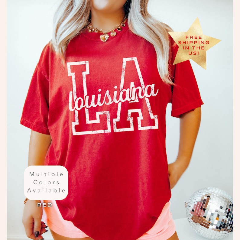 May include: A red t-shirt with the text "LA Louisiana" in white. The shirt has a vintage look and is available in multiple colors. A gold star graphic with text is in the upper right corner.
