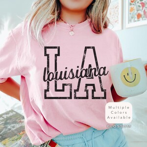 May include: A light pink sweatshirt with the text "LA Louisiana" in black. The letters "LA" are in a block font, and "Louisiana" is in a script font. The image also includes a white mug with a yellow smiley face.
