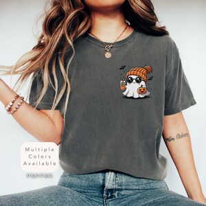 Pode incluir: Uma camiseta cinza com um fantasma de desenho animado usando um gorro e &oacute;culos de sol, segurando uma ab&oacute;bora e uma x&iacute;cara. O texto "Multiple Colors Available" e "PEPPER" est&atilde;o na camiseta.