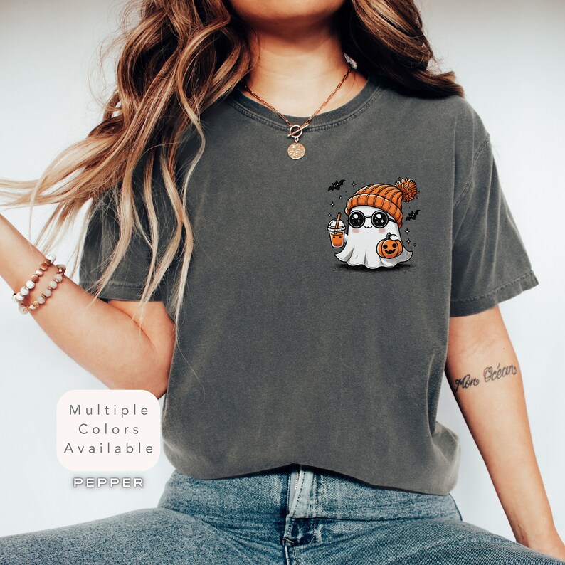 Pode incluir: Uma camiseta cinza com um fantasma de desenho animado usando um gorro e &oacute;culos de sol, segurando uma ab&oacute;bora e uma x&iacute;cara. O texto "Multiple Colors Available" e "PEPPER" est&atilde;o na camiseta.