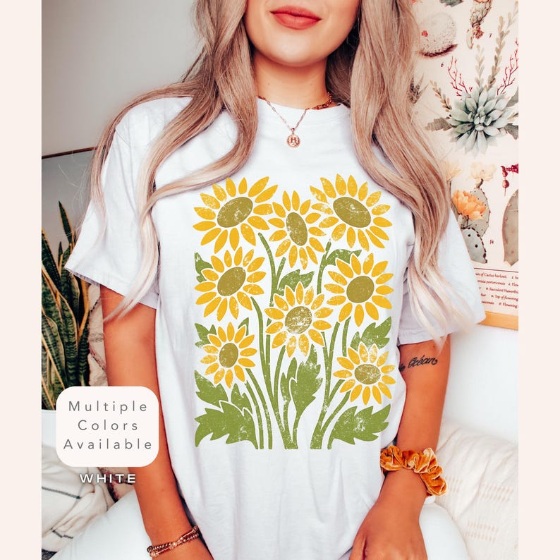 May include: White t-shirt with a vintage-style graphic of yellow sunflowers with green leaves. The text "Multiple Colors Available" and "WHITE" is printed on the shirt.