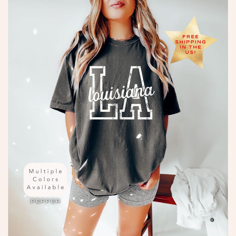May include: Dark gray t-shirt with the text "LA Louisiana" in white, distressed lettering. The shirt has a vintage, worn look. The image also includes a gold star with the text "FREE SHIPPING IN THE US!"