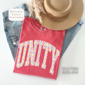 Può includere: Una maglietta rossa con la scritta "UNITY" in un motivo a scacchi, un cappello color cachi e jeans blu. La maglietta è etichettata "WATERMELON" e "Multiple Colors Available". La maglietta è piegata sopra i jeans.