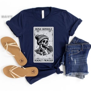 May include: A navy blue t-shirt with a graphic of a skeleton wearing a beanie and eating a pickle. The graphic is in a card-like frame with the text "Dead Serious About Pickles".