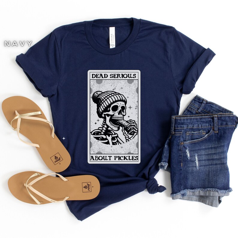 May include: A navy blue t-shirt with a graphic of a skeleton wearing a beanie and eating a pickle. The graphic is in a card-like frame with the text "Dead Serious About Pickles".