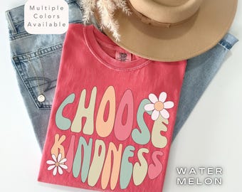 Puede incluir: Una camiseta roja con las palabras "CHOOSE KINDNESS" en una fuente retro, con gráficos de flores. La camiseta se combina con unos vaqueros de lavado claro y un sombrero color canela. También es visible el texto "WATER MELON".