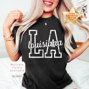 May include: Black t-shirt with the text "LA Louisiana" in white. The letters "LA" are in a block letter style, with "Louisiana" written in a cursive font. The shirt is part of a collection with multiple colors available.