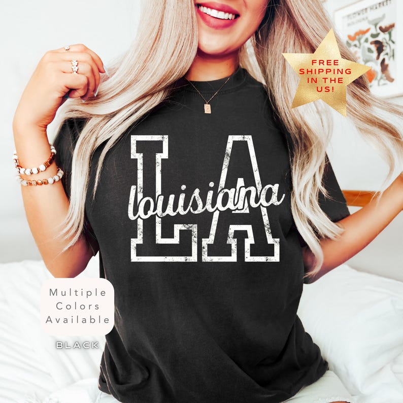 May include: Black t-shirt with the text "LA Louisiana" in white. The letters "LA" are in a block letter style, with "Louisiana" written in a cursive font. The shirt is part of a collection with multiple colors available.