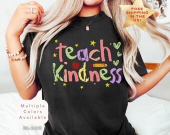 May include: Black t-shirt with the words "Teach Kindness" in colorful letters, decorated with stars, hearts, and a pencil. The shirt also has the text "Multiple Colors Available" and "BLACK".