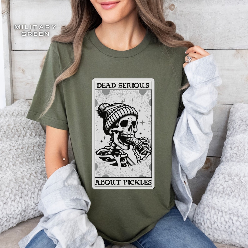 May include: A green t-shirt with a graphic of a skeleton wearing a beanie and eating a pickle. The graphic is in a card-like frame with the text "Dead Serious About Pickles".