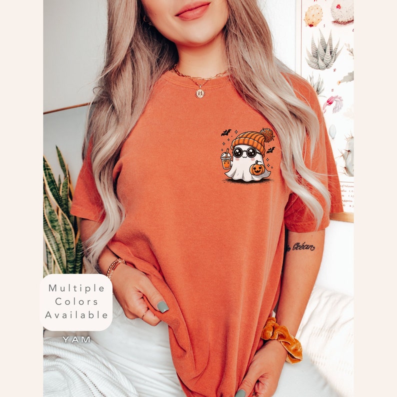 Pode incluir: Uma camiseta laranja queimada com um fantasma de desenho animado fofo usando um gorro e &oacute;culos de sol, segurando uma ab&oacute;bora e uma bebida. O texto "Multiple Colors Available" est&aacute; na parte inferior esquerda da imagem.