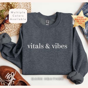 May include: Dark heather grey sweatshirt with the text "vitals & vibes" printed in white.