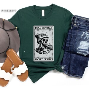May include: A green t-shirt with a graphic of a skeleton wearing a beanie and eating a pickle. The graphic is in a card-like frame with the text "Dead Serious About Pickles".