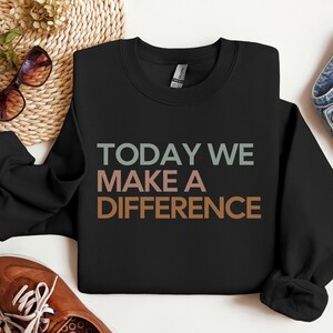 Puede incluir: Sudadera negra con el texto "TODAY WE MAKE A DIFFERENCE" en un degradado de verde, marrón y naranja.