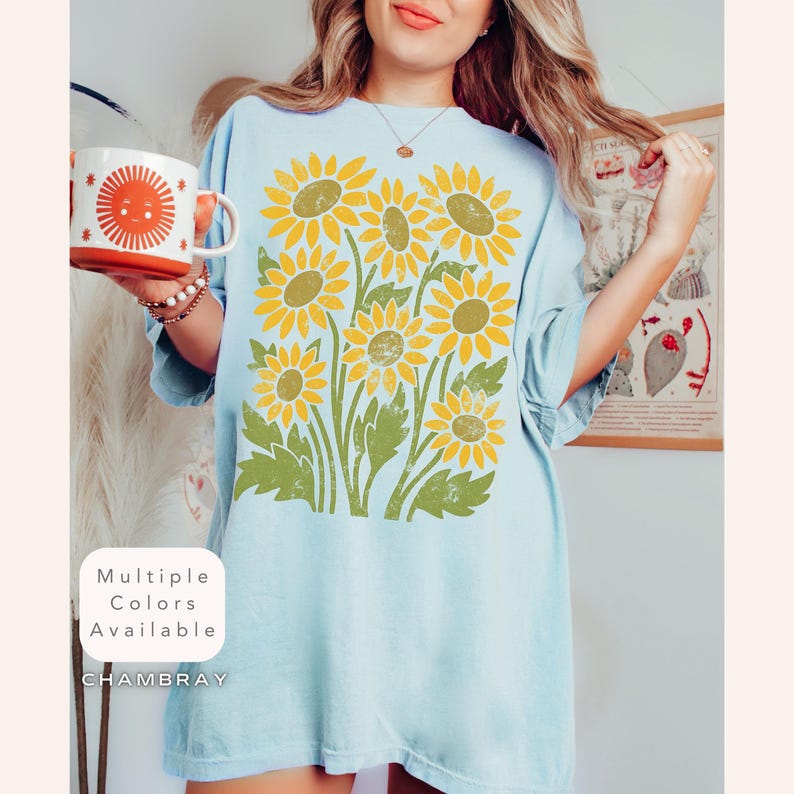May include: A light blue chambray t-shirt with a vintage-style graphic of yellow sunflowers. The text "Multiple Colors Available" and "CHAMBRAY" are printed on the shirt.