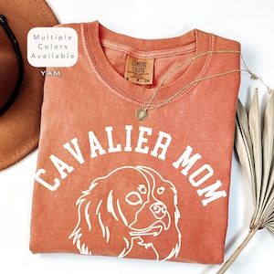 Peut inclure: Un t-shirt couleur terre cuite avec les mots "CAVALIER MOM" en lettres blanches et un dessin blanc d'un Cavalier King Charles Spaniel. Le t-shirt a un col rond et un collier dor&eacute;. Le texte "Multiple Colors Available" est dans un encadr&eacute; blanc.