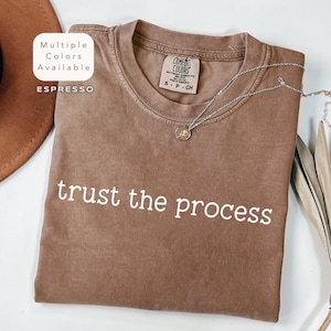 May include: A brown t-shirt with the text "trust the process" printed in white. The shirt is made of a soft, comfortable material and has a relaxed fit.