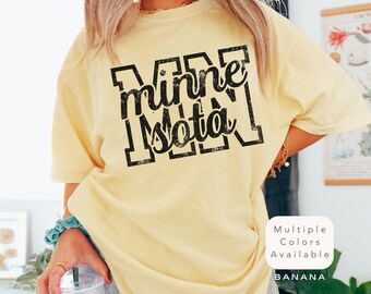 Op de afbeelding: Een lichtgeel t-shirt met het woord "Minnesota" in een zwarte, verweerde letter. Het shirt heeft een relaxte pasvorm en korte mouwen. De tekst "Multiple Colors Available" en de kleur "Banana" zijn ook zichtbaar.