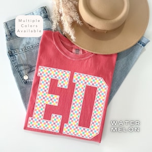 May include: A red t-shirt featuring the letters "ED" in a colourful chequered design. The shirt is folded on top of blue jeans and a tan hat. The text "Watermelon" is visible on the bottom right.