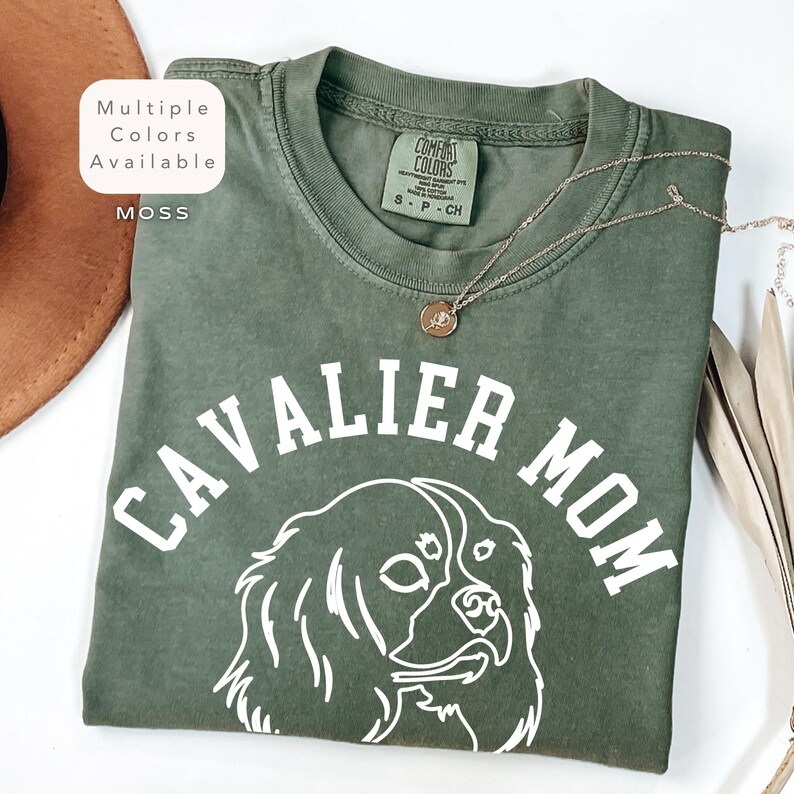 Peut inclure: T-shirt vert mousse avec l'inscription blanche "CAVALIER MOM" et le contour blanc d'un chien Cavalier King Charles Spaniel. Le t-shirt a un col rond et des manches courtes. Un collier dor&eacute; avec un pendentif est pos&eacute; sur le t-shirt.