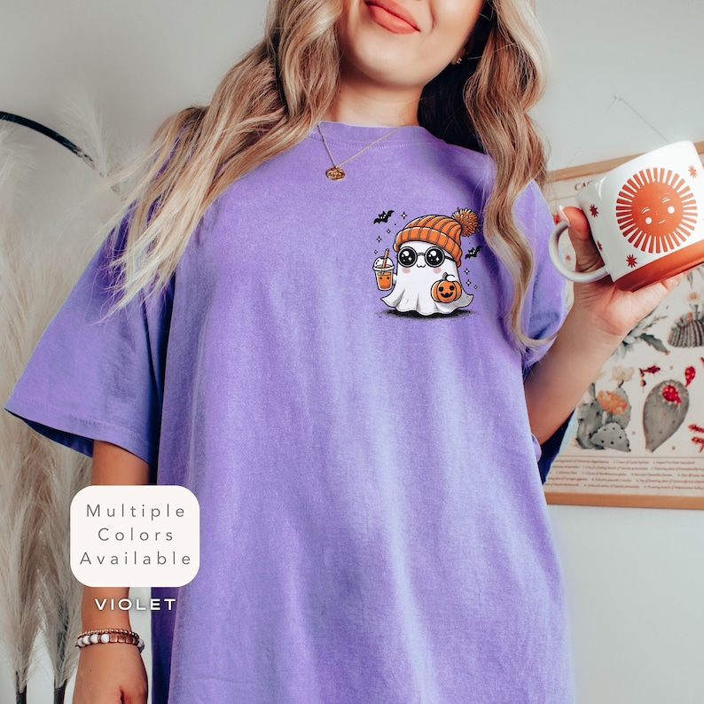 Pode incluir: Uma camiseta roxa com um fantasma de desenho animado usando um gorro, &oacute;culos de sol e segurando uma ab&oacute;bora e uma bebida. O texto "Multiple Colors Available" e "VIOLET" est&aacute; impresso na camiseta.
