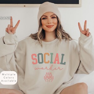 Könnte beinhalten: Ein beiger Pullover mit dem Text "SOCIAL worker" in Regenbogenfarben. Der Text ist von einem rosa Umriss umgeben. Unter dem Text befindet sich ein rosa Symbol von zwei Händen, die drei Personen halten.