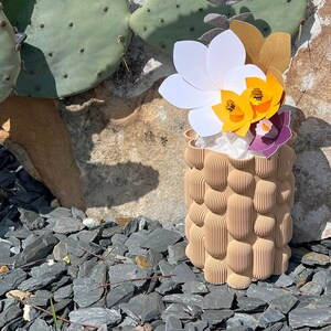 May include: A white, yellow, and purple paper flower arrangement in a textured brown ceramic vase. The vase is sitting on a bed of gray rocks.