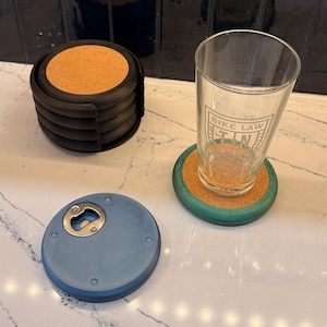 May include: A set of black coasters with cork centers, a blue bottle opener, and a glass with the text "BIKE LAW" printed on it. A green coaster with a cork center is under the glass.
