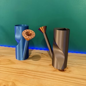 May include: Two decorative watering cans. One is blue with a copper-colored spout, and the other is gray with a copper-colored spout. The watering cans have a unique, twisted design. The background is a teal color.