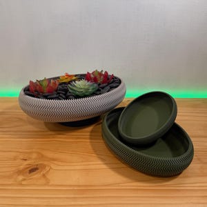 May include: An oval-shaped planter with a white and gray zigzag pattern, filled with black stones and colorful faux succulents. Two olive green oval dishes with a matching zigzag pattern are next to the planter.