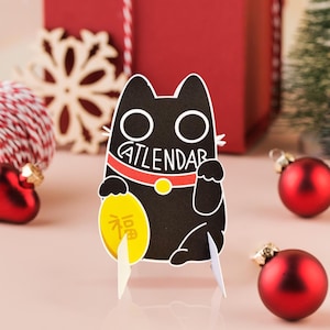 Puede incluir: Un calendario de papel con forma de gato negro con la palabra "CATLENDAR" impresa en el collar. El gato sostiene una moneda de oro con caracteres chinos. El calendario está rodeado de adornos navideños rojos.