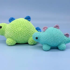 May include: Two crocheted dinosaur toys. The larger dinosaur is green with blue accents and the smaller dinosaur is blue with purple accents.