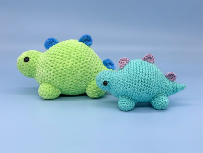 May include: Two crocheted dinosaur toys. The larger dinosaur is green with blue accents and the smaller dinosaur is blue with purple accents.