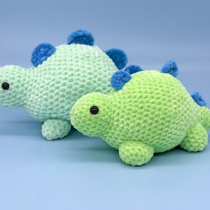 May include: Two crocheted dinosaur toys, one green and one light blue, with blue accents. The dinosaurs are sitting on a light blue background.