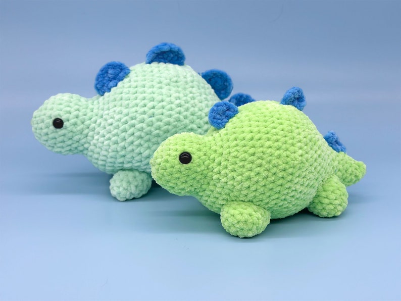 May include: Two crocheted dinosaur toys, one green and one light blue, with blue accents. The dinosaurs are sitting on a light blue background.