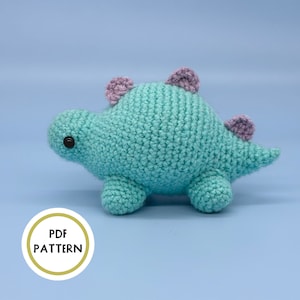 May include: A crocheted turquoise dinosaur toy with purple ears. The toy has black eyes and a small tail.