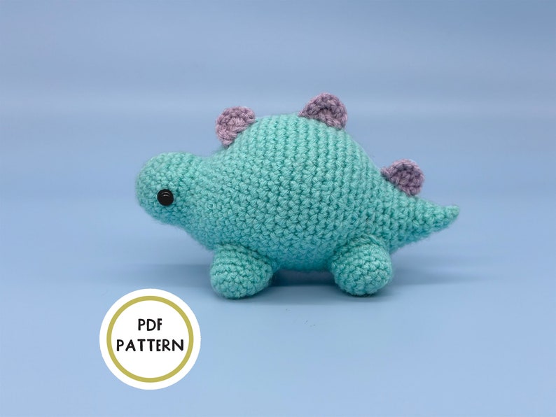 May include: A crocheted turquoise dinosaur toy with purple ears. The toy has black eyes and a small tail.